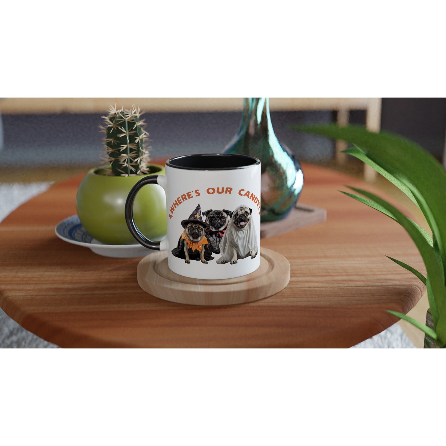 Round wooden end table with a potted cactus, glass vase, and a white mug on it. The mug has a black handle, rim, and inside with a picture of three pugs--one dressed as a witch, one dressed as a vampire, and one dressed as a ghost with the words "Where's our candy?" arched over them.
