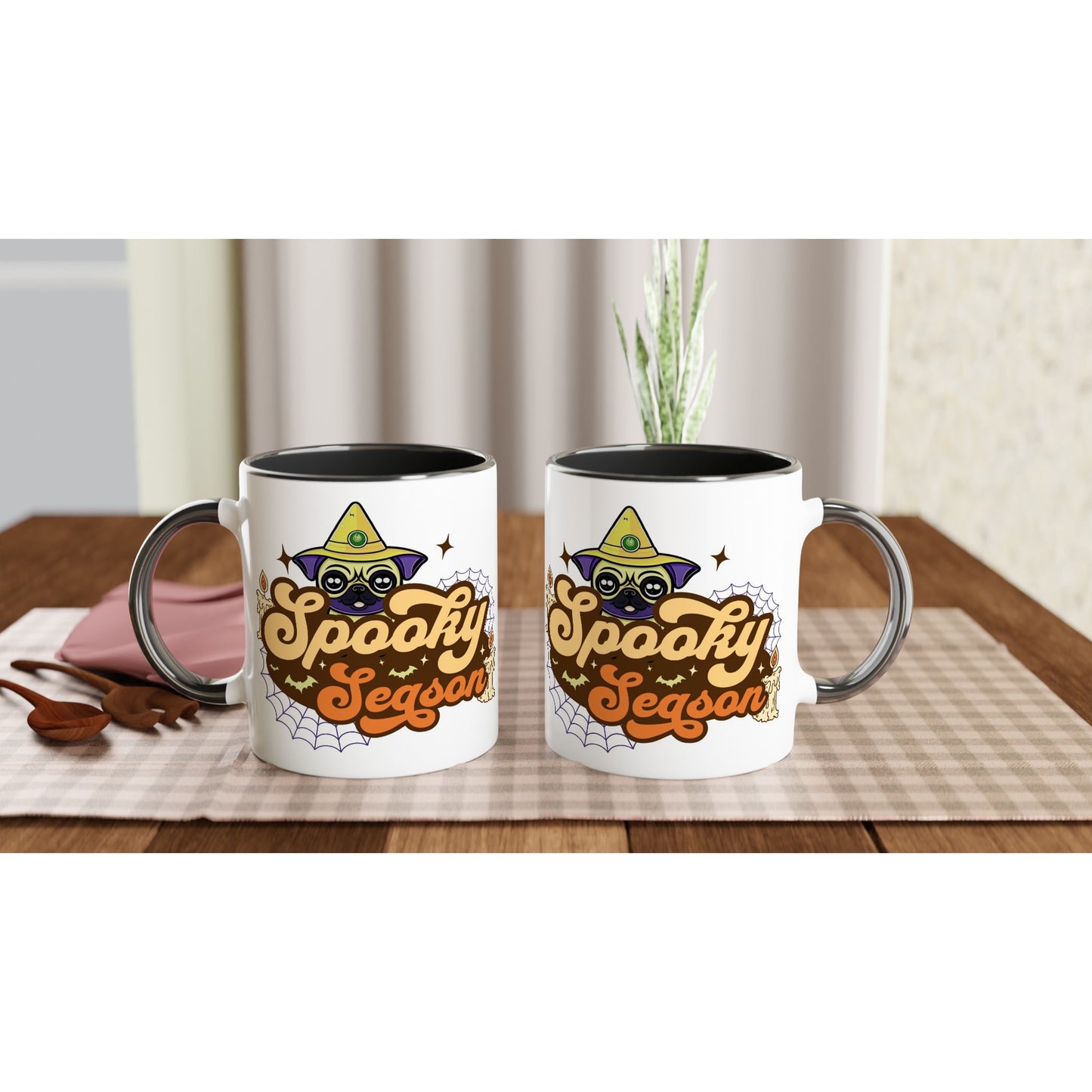 Two white mugs with black handles, rims, and insides, each facing a different direction, with the image of a pug's head in a witch's hat and the words "Spooky Season" below it. The image also has cobwebs, bats, and candles in it. The mugs are sitting on a wooden kitchen table on a gray checkered placemat next to a napkin, a wooden fork, and a wooden spoon.