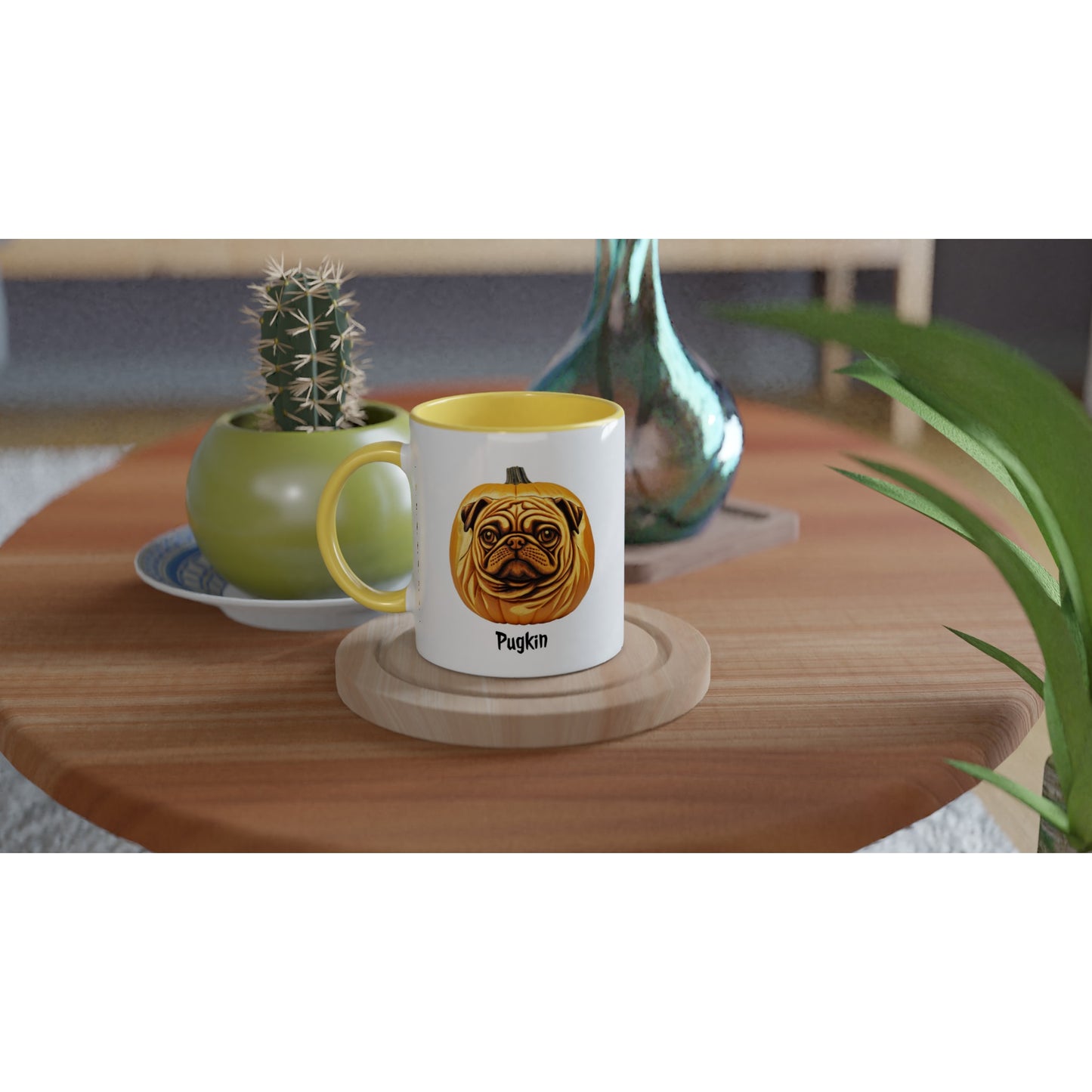 round wooden end table with a potted cactus, glass vase, and white  mug on it. The mug has a yellow handle, rim, and inside and has the image of a pumpkin that has the face of a pug on it. The word Pugkin is written beneath the image.