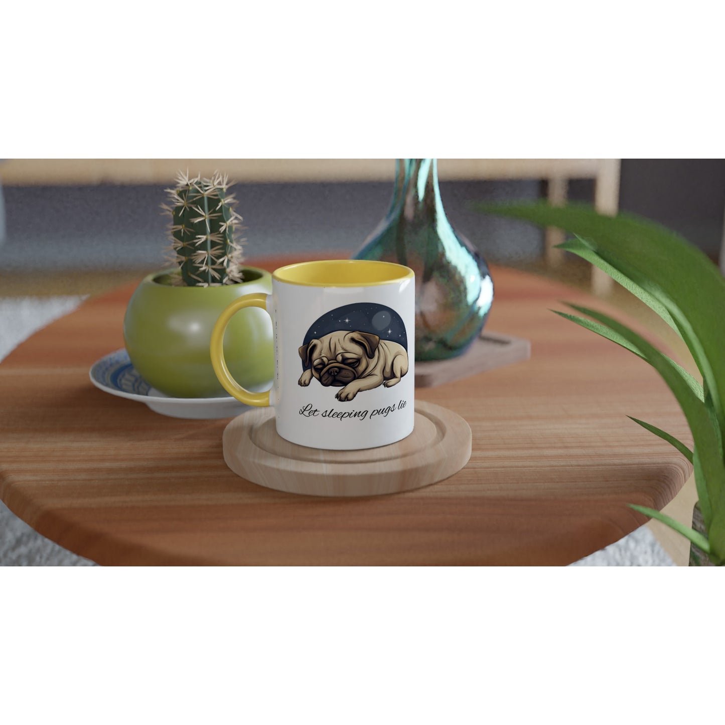 Round wooden end table with a potted cactus, glass vase, and white mug on it. The mug has a yellow handle, rim, and inside and has a cartoonish picture of a sleeping pug surrounded by a half-circle of starry night sky with the words "Let sleeping pugs lie" under the image in cursive lettering.