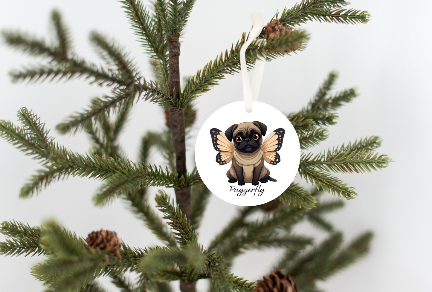 3 inch round white ornament hanging in a Christmas tree.  The ornament has a cartoon style picture of a fawn-colored pug with orangey-beige butterfly wings attached to her back and big sad eyes. The word "Puggerfly" is beneath the image.