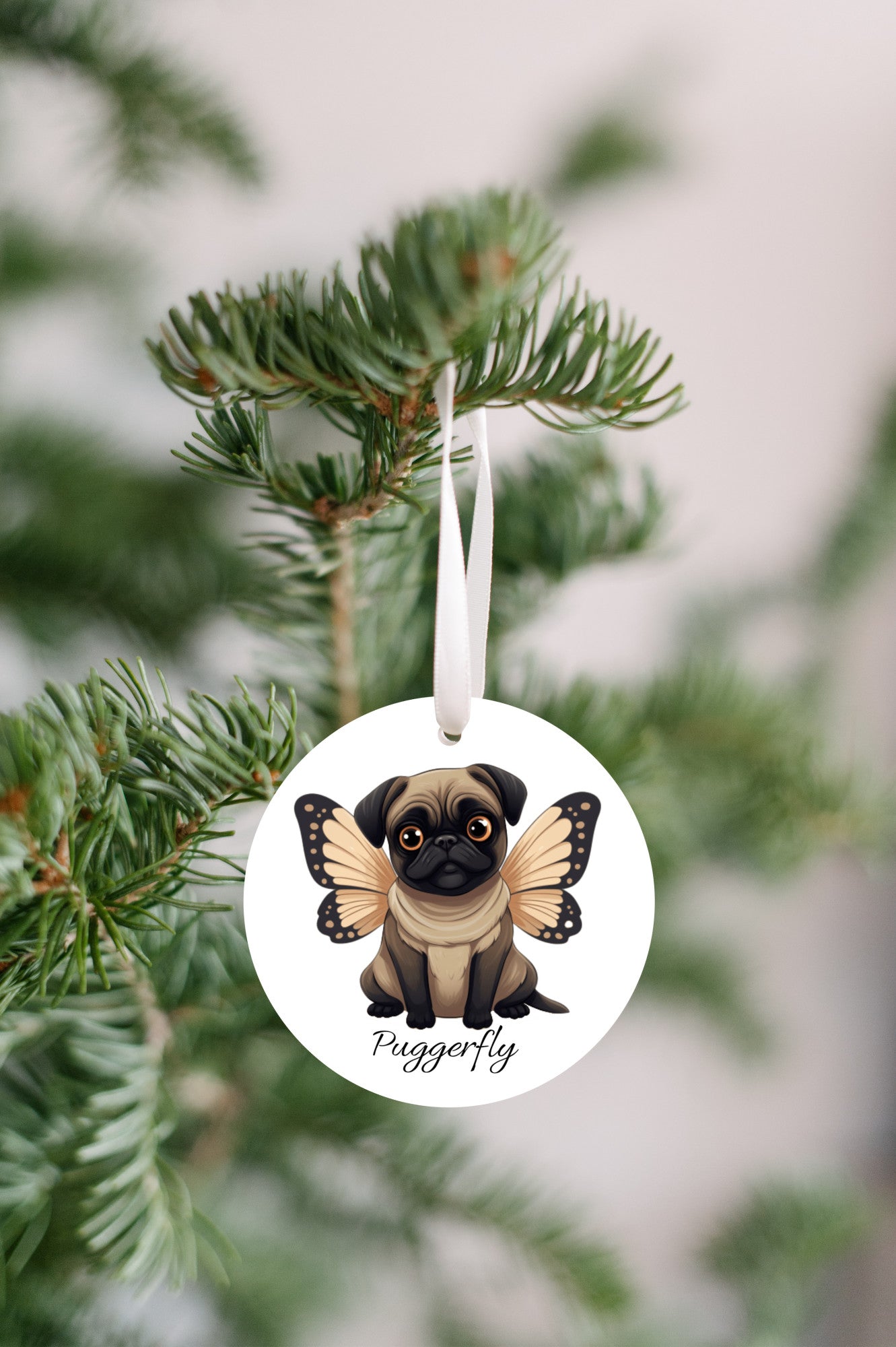 3 inch round white ornament hanging in a Christmas tree.  The ornament has a cartoon style picture of a fawn-colored pug with orangey-beige butterfly wings attached to her back and big sad eyes. The word "Puggerfly" is beneath the image.