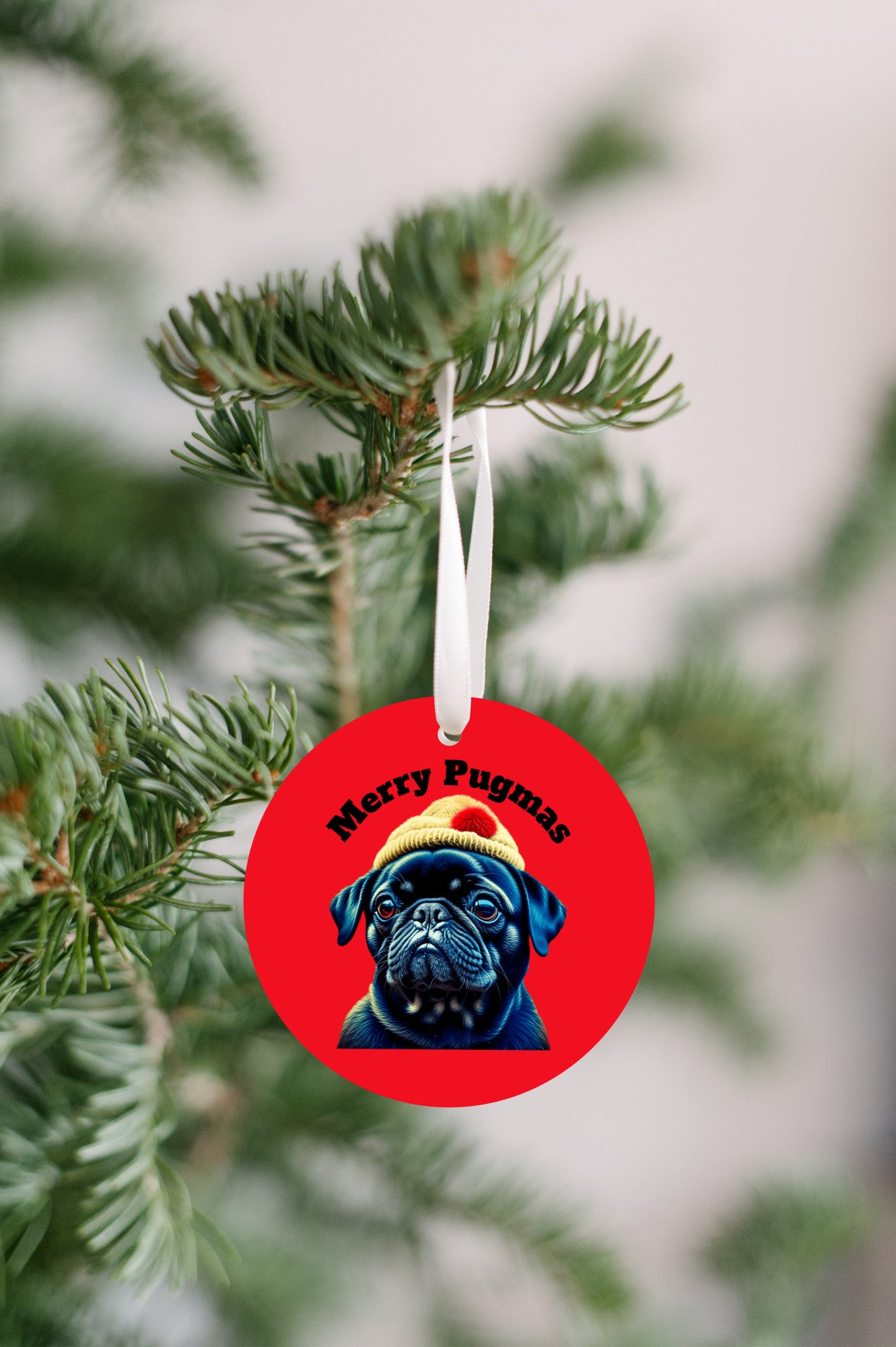 3 inch round ornament hanging from a Christmas tree. The ornament is red with the words "Merry Pugmas" arched over the head of a black pug who is wearing a yellow winter hat with a pompom on it.