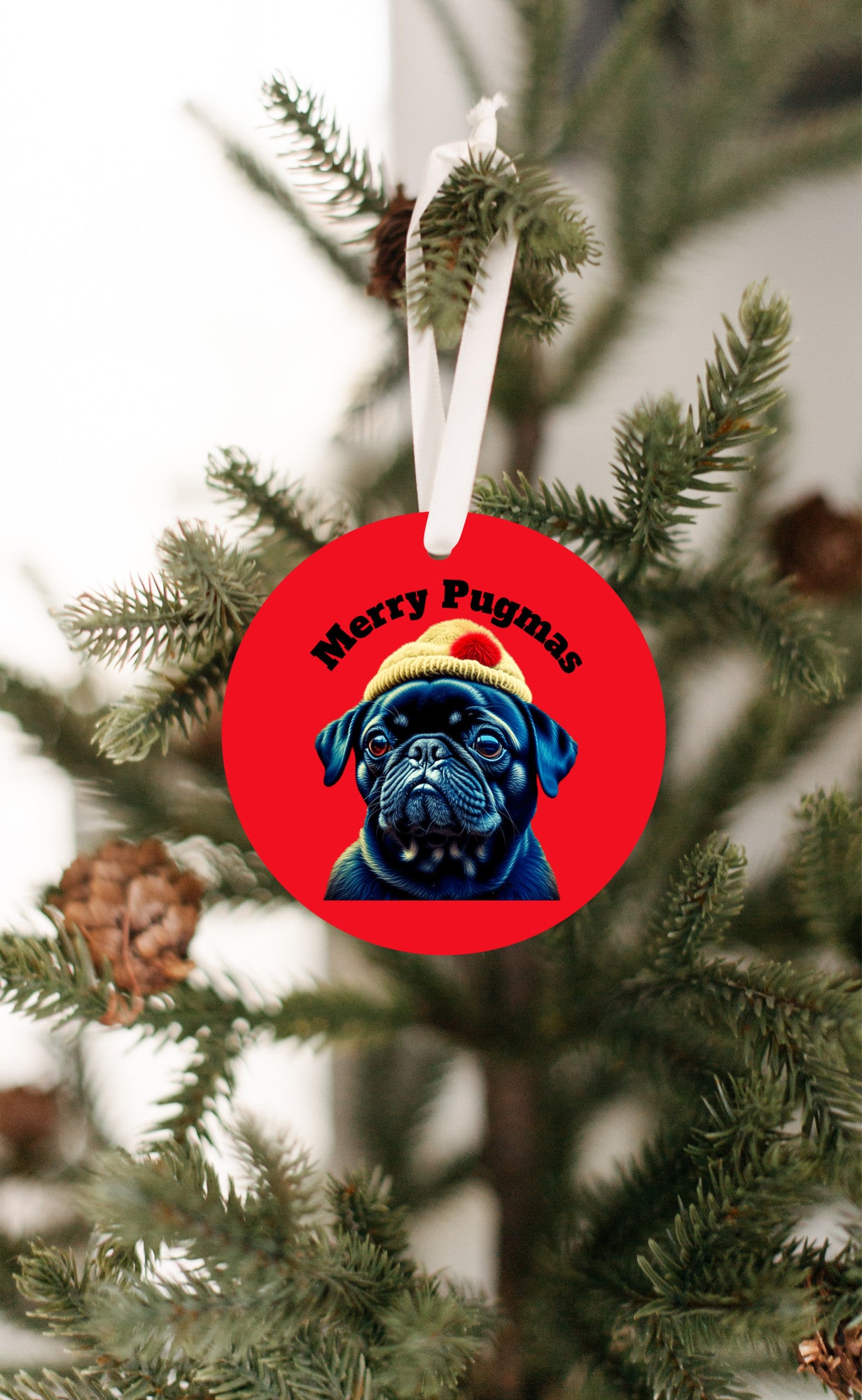 3 inch round ornament hanging from a Christmas tree. The ornament is red with the words "Merry Pugmas" arched over the head of a black pug who is wearing a yellow winter hat with a pompom on it.