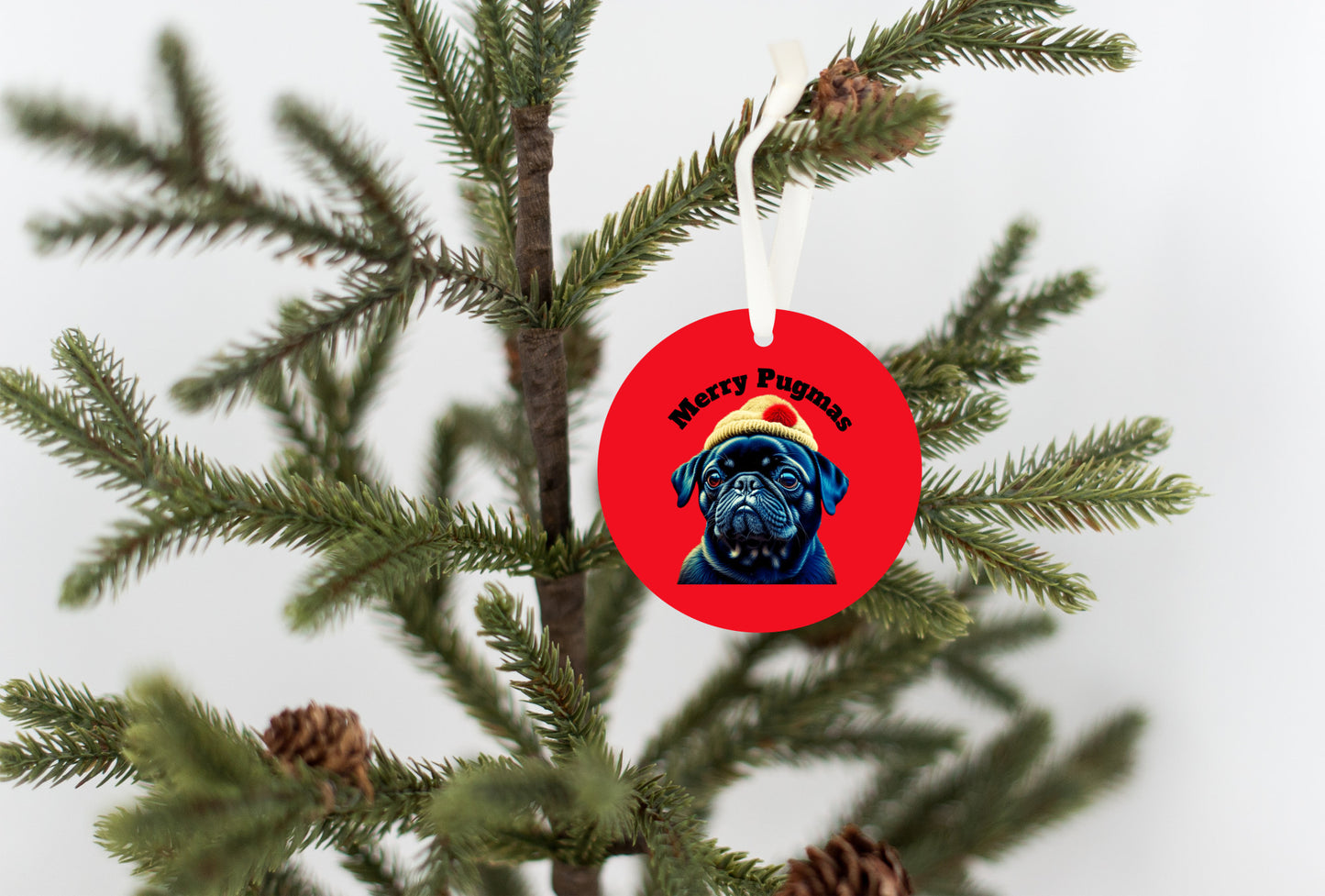 3 inch round ornament hanging from a Christmas tree. The ornament is red with the words "Merry Pugmas" arched over the head of a black pug who is wearing a yellow winter hat with a pompom on it.