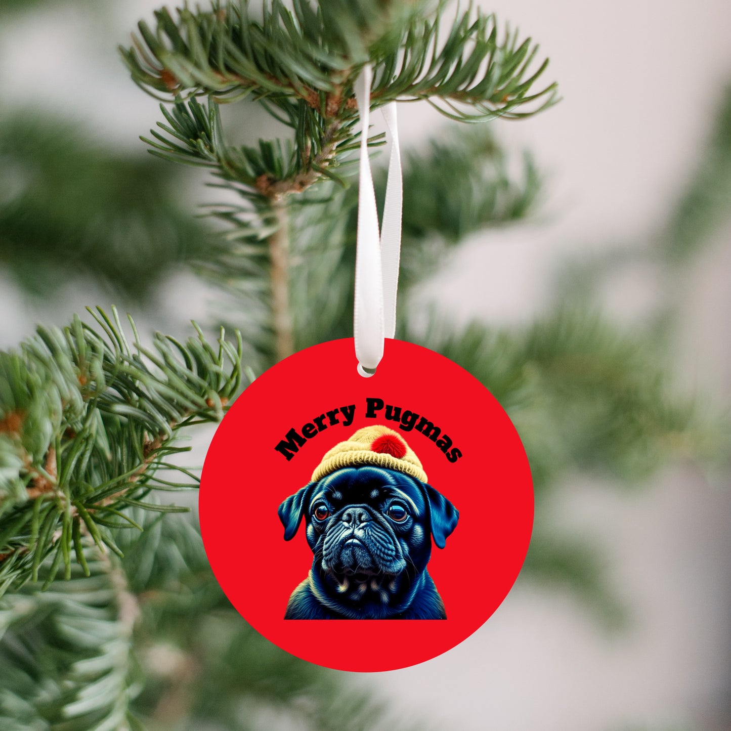 3 inch round ornament hanging from a Christmas tree. The ornament is red with the words "Merry Pugmas" arched over the head of a black pug who is wearing a yellow winter hat with a pompom on it.