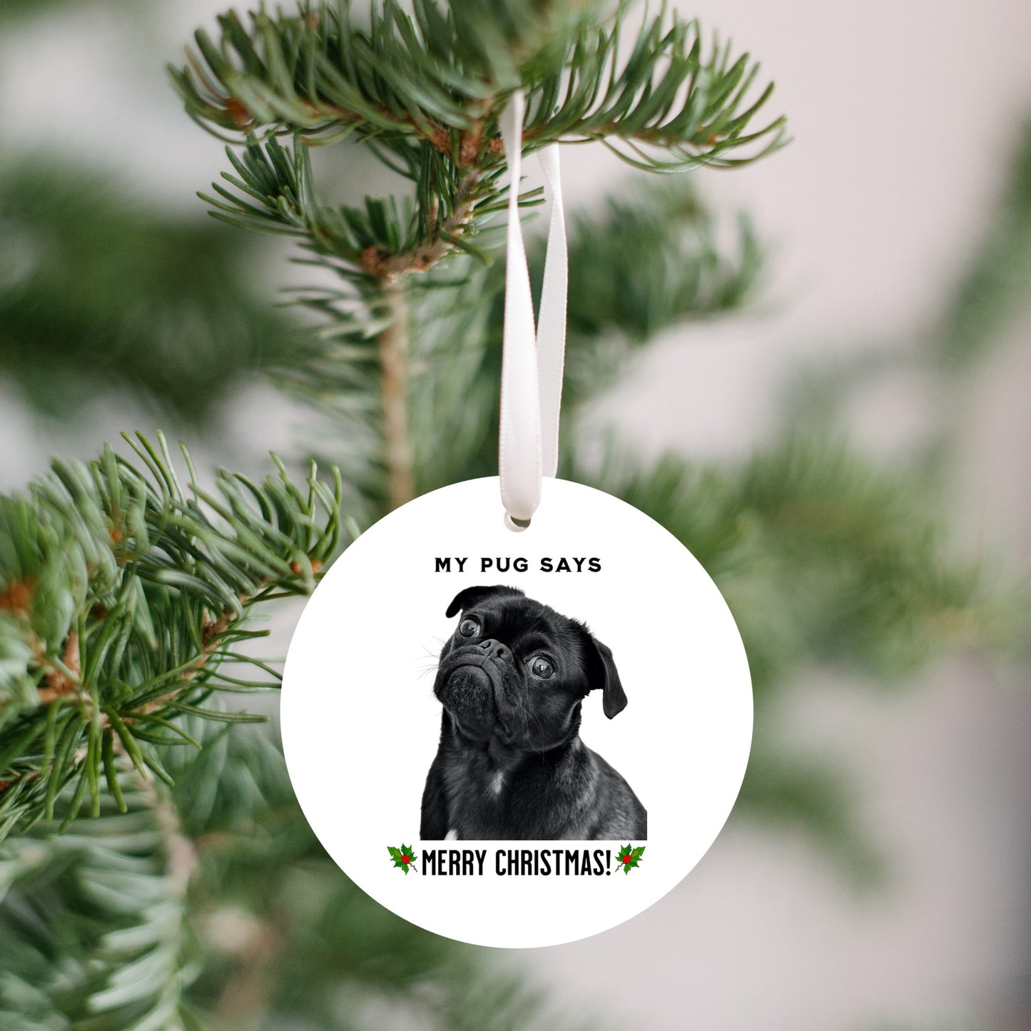 3 inch round flat white ornament hanging in a Christmas tree with a picture of the head and shoulders of a black pug with the words "My pug says" above the pug and "Merry Christmas" below the pug. 