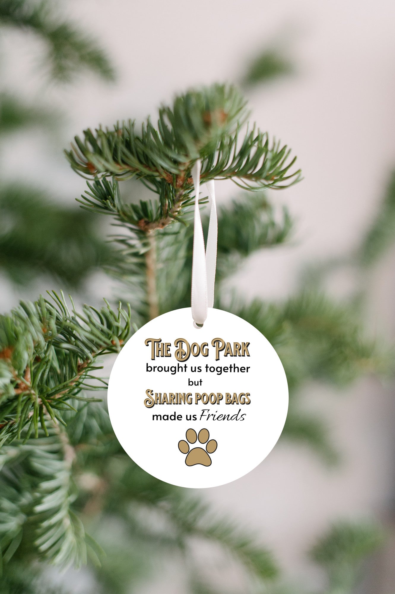 3 inch round flat white ornament hanging in a Christmas tree. On the ornament are the words "The Dog Park brought us together but sharing poop bags made us friends" in black and gold letters. There is a gold pawprint beneath the words.