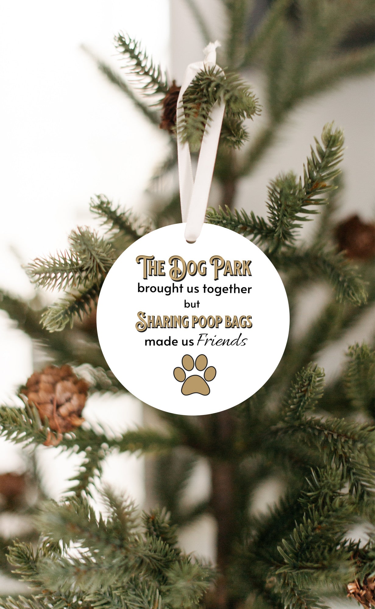 3 inch round flat white ornament hanging in a Christmas tree. On the ornament are the words "The Dog Park brought us together but sharing poop bags made us friends" in black and gold letters. There is a gold pawprint beneath the words.