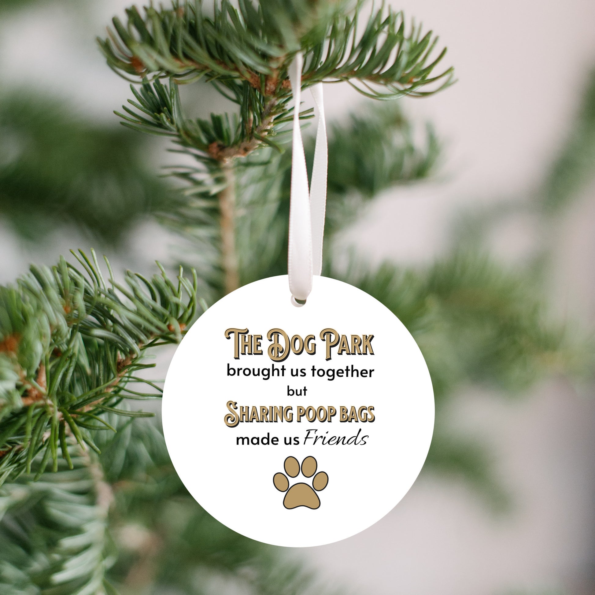 3 inch round flat white ornament hanging in a Christmas tree. On the ornament are the words "The Dog Park brought us together but sharing poop bags made us friends" in black and gold letters. There is a gold pawprint beneath the words.